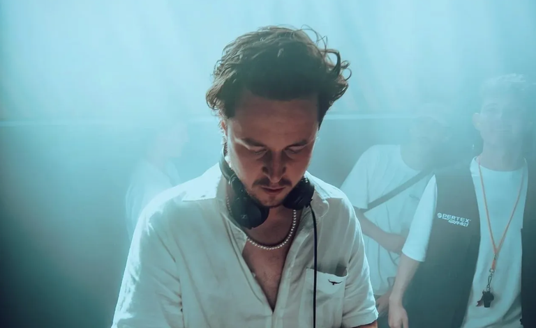 DJ Jake Phillips performing live on stage with headphones around his neck in a atmospheric, hazy promotional photo.